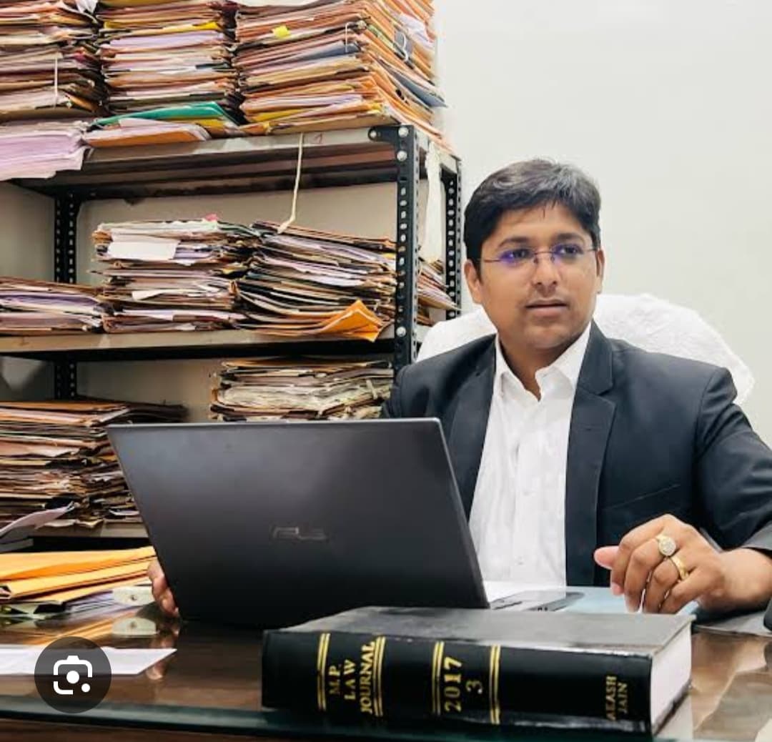 Man in a suit sitting at a desk with a laptop and stacks of legal files.