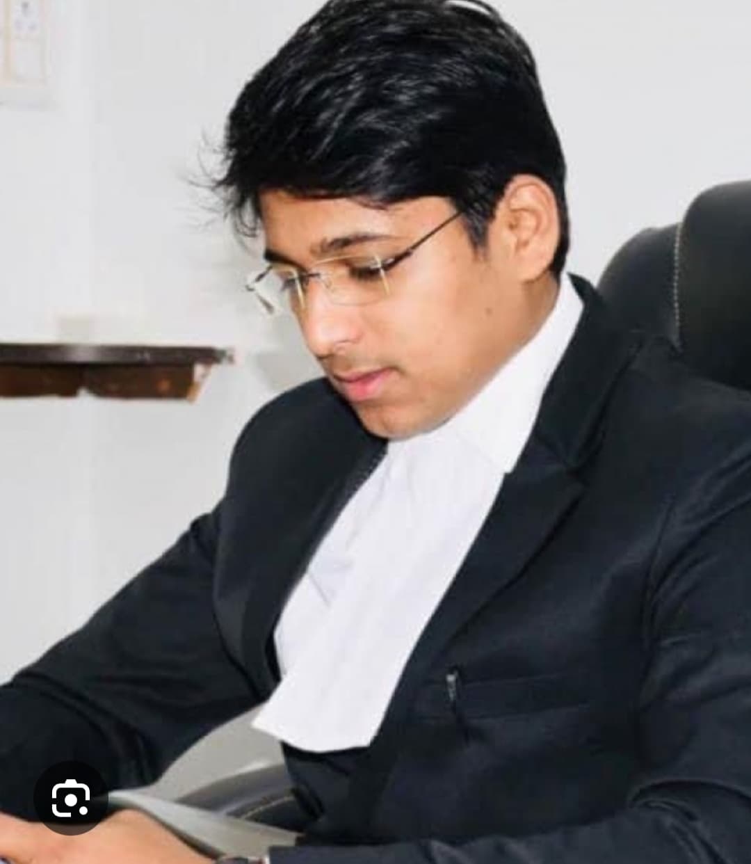 Young man in a black suit and white legal neckband looking down while sitting.