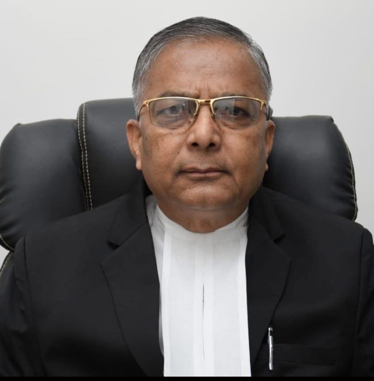 Serious man with glasses wearing a black blazer and white legal neckband in a chair.