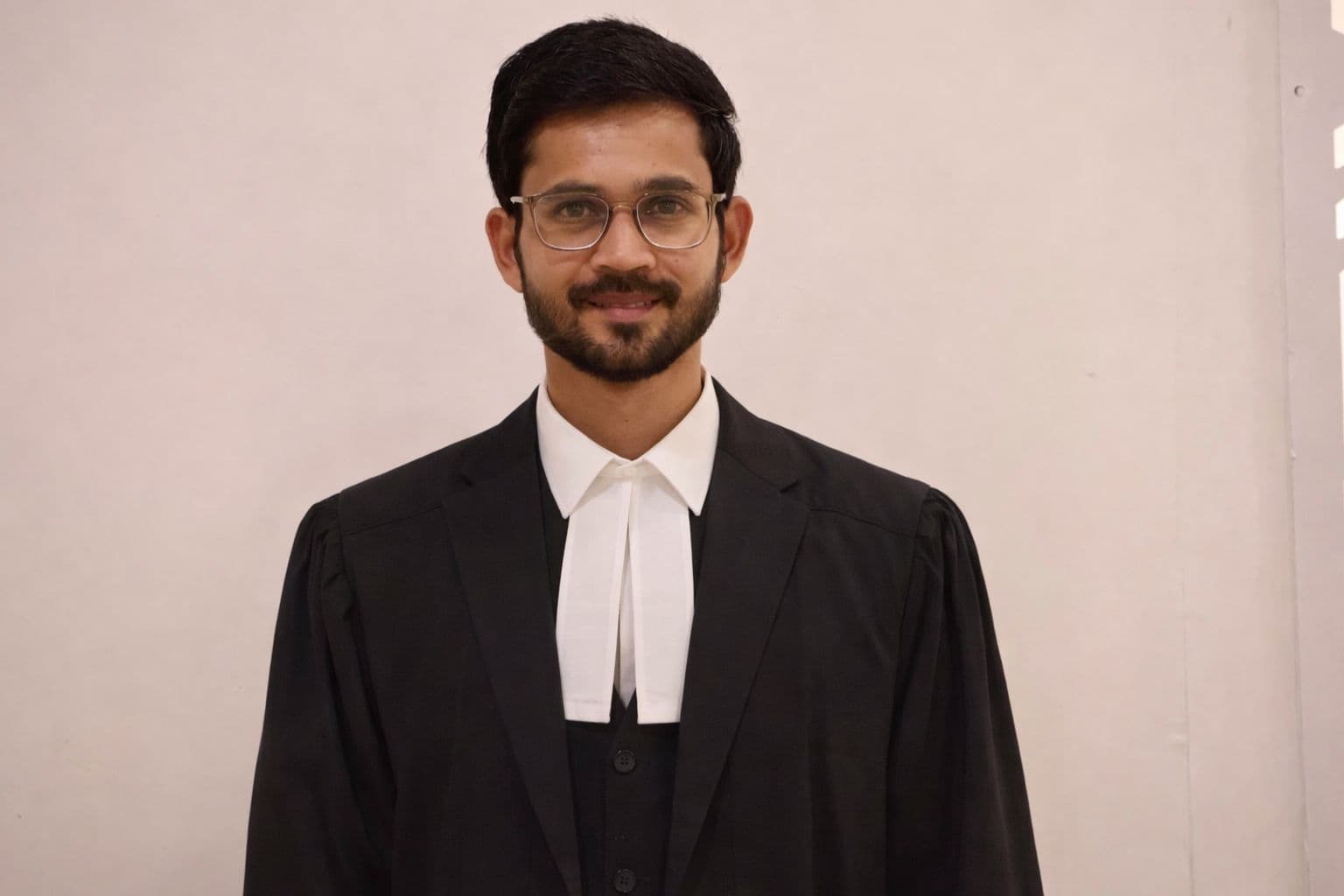 A bearded man wearing glasses and a black legal gown with white neckbands, smiling.