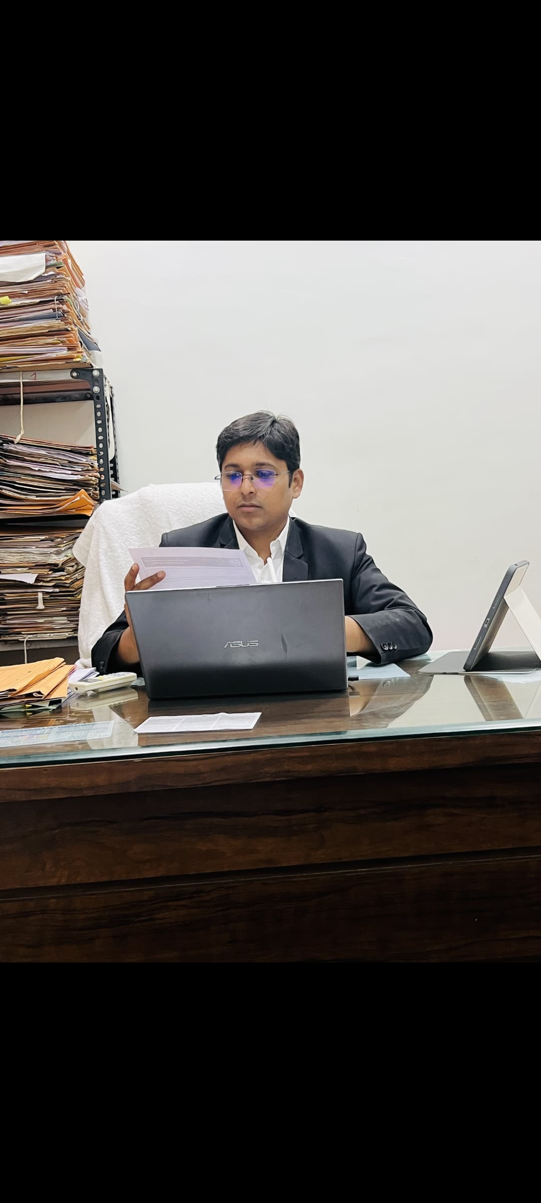 Man in a suit and glasses reviews documents at a desk with file stacks.
